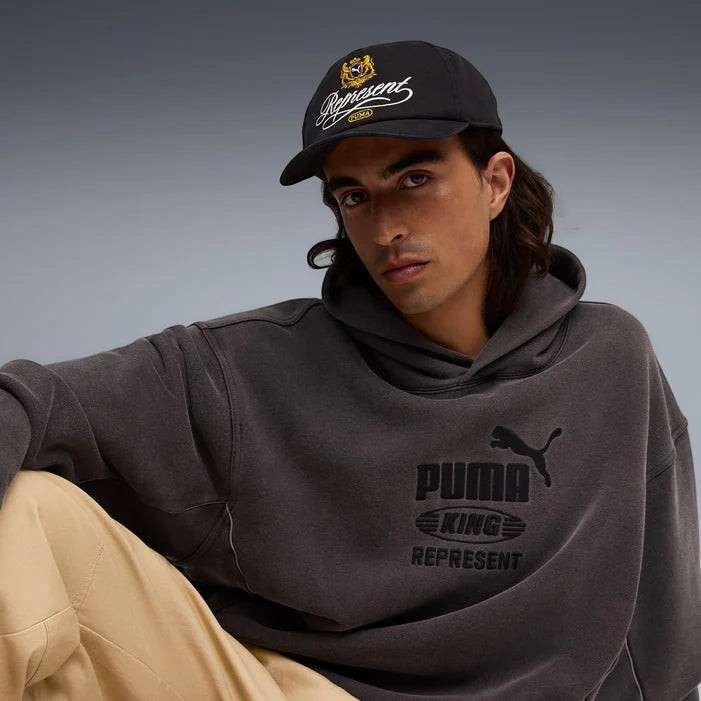 Drop PUMA x REPRESENT black baseball cap with gold and white embroidered logo worn by a man in a grey hoodie
