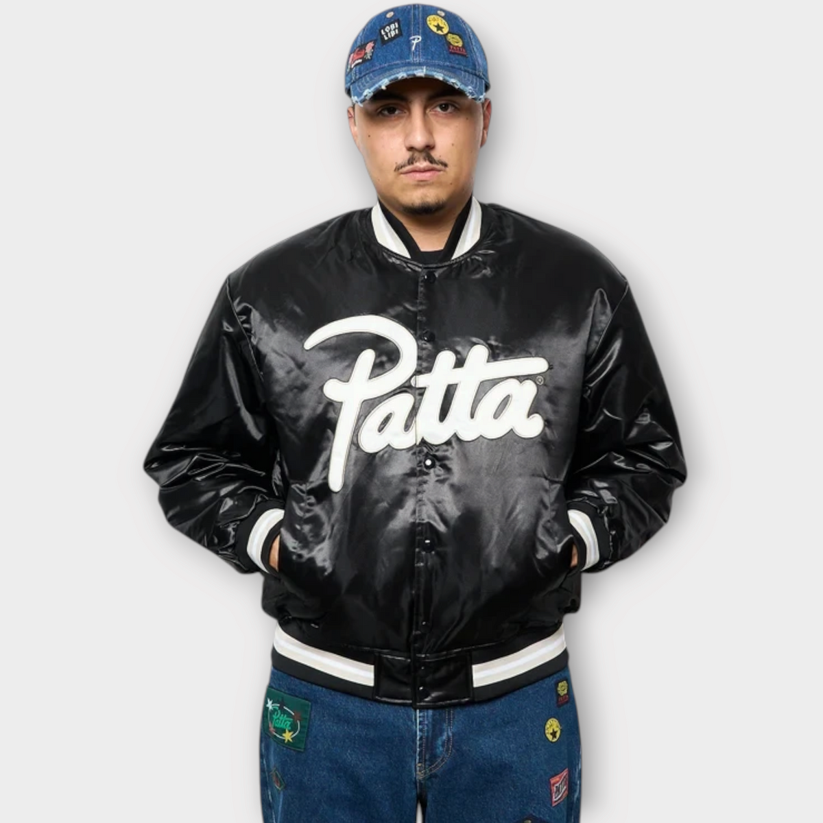 Drop Patta black satin varsity jacket with white script logo, worn by a man in a denim cap and jeans