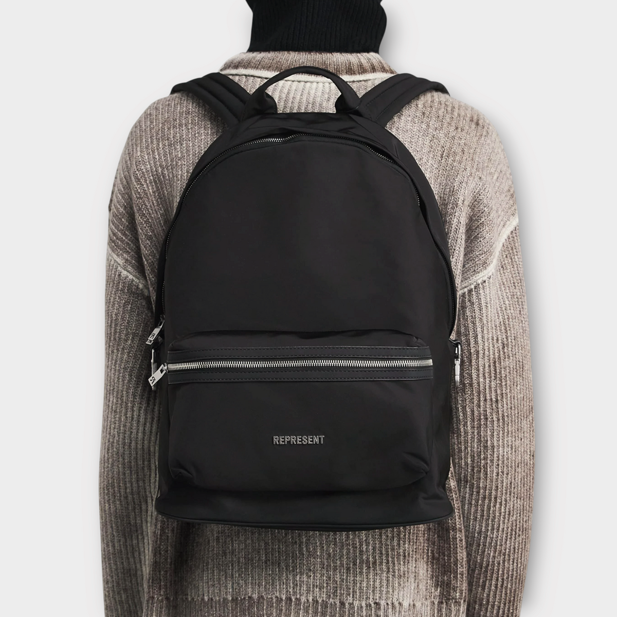 Drop Represent black nylon backpack with silver zipper details worn on the back over a textured sweater